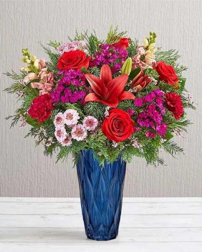 <p>Celebrate the holidays with an arrangement as rich and refined as the season itself. The Yuletide Jewel Bouquet brings timeless elegance to winter celebrations, combining dramatic colors, lush textures, and heirloom-inspired style. Whether used as a centerpiece or sent as a meaningful gift, this luxurious bouquet is a striking expression of classic holiday beauty.</p>
<p>This festive design features a stunning mix of jewel-toned blooms, thoughtfully selected to evoke Victorian grandeur. Deep red roses, velvety carnations, and other richly colored seasonal flowers are paired with elegant greenery to create a bouquet full of depth, contrast, and visual drama. The flowers are hand-arranged in a tall, ribbed sapphire-blue glass vase—adding bold color and artisanal character to the overall presentation.</p>
<p>Ideal for Christmas gatherings, holiday dinner parties, or as a gift for someone who loves traditional décor, the Yuletide Jewel Bouquet brings warmth, luxury, and sophistication to any space. The classic combination of jewel tones and festive greenery makes it perfect for mantels, dining tables, or entryway displays.</p>
<p>Each arrangement is handcrafted by expert local florists using the freshest seasonal blooms. Some flowers may arrive in bud form for longer-lasting enjoyment. Local delivery is available, ensuring your gift arrives fresh, beautifully styled, and ready to impress.</p>
<p>Send the Yuletide Jewel Bouquet today and let its rich colors and timeless design bring holiday magic to someone’s home.</p>