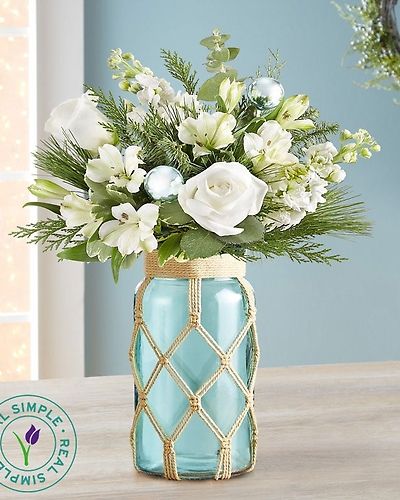 <p>Bring the breezy charm of the beach to your holiday celebration with this coastal-inspired bouquet—a fresh and unexpected take on Christmas for those who dream of ocean waves even in winter. With its serene palette and artistic details, this design beautifully blends seasonal cheer with seaside calm.</p>
<p>This airy arrangement features soft white roses and carnations, accented by fragrant evergreens and playful turquoise ornaments that shimmer like sea glass. The blooms are arranged in a sea-green glass jar reminiscent of recycled beach glass, wrapped in a handmade macramé cover for a touch of eco-conscious, Boho flair. More than just a vase, the container becomes a lasting piece of décor with its coastal, minimalist charm.</p>
<p>Ideal for holiday gifting or decorating, this bouquet is perfect for warm-weather celebrations, beach houses, or anyone who prefers palm trees to pine trees. It’s a thoughtful choice for friends, family, or coworkers who love relaxed, modern style with a natural touch. Whether placed on a kitchen counter, office desk, or holiday table, it radiates peace, joy, and coastal elegance.</p>
<p>Each arrangement is handcrafted using the freshest seasonal blooms, and some flowers may arrive in bud form to ensure long-lasting beauty. Local delivery is available through our trusted florist network, offering a seamless way to share holiday cheer with care and convenience.</p>
<p>With its refreshing scent, soothing tones, and artistic presentation, the Seaside Celebration bouquet adds a little escape—and a lot of style—to the season.</p>
<p>Send a splash of coastal serenity this holiday with a bouquet that brings the beach to Christmas in the most beautiful way.</p>